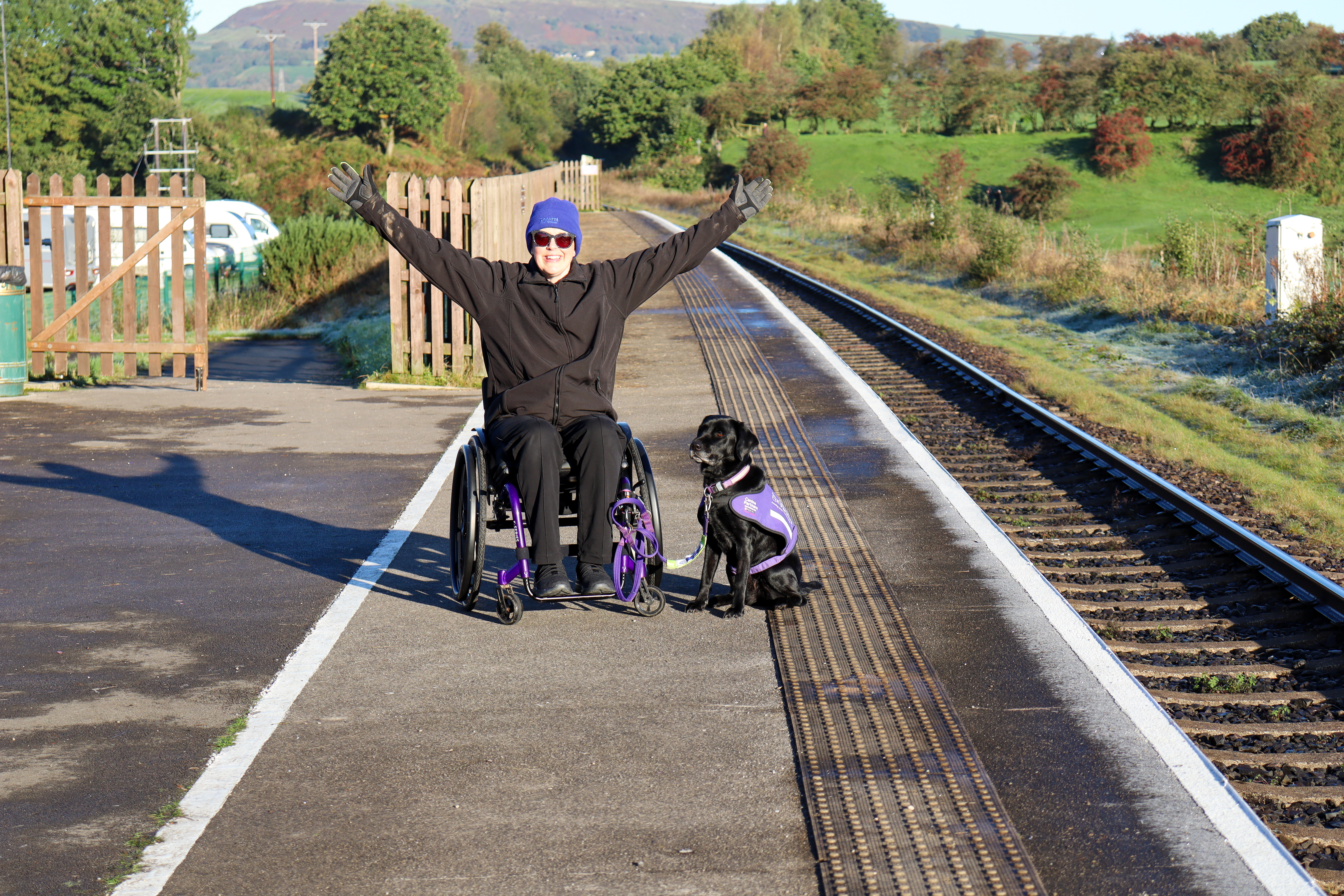 Burrs Country Park railway platform which is beautifully clean and tidy. Nicki is in her wheelchair looking excited, with her assistance dog, Liggy beside her.