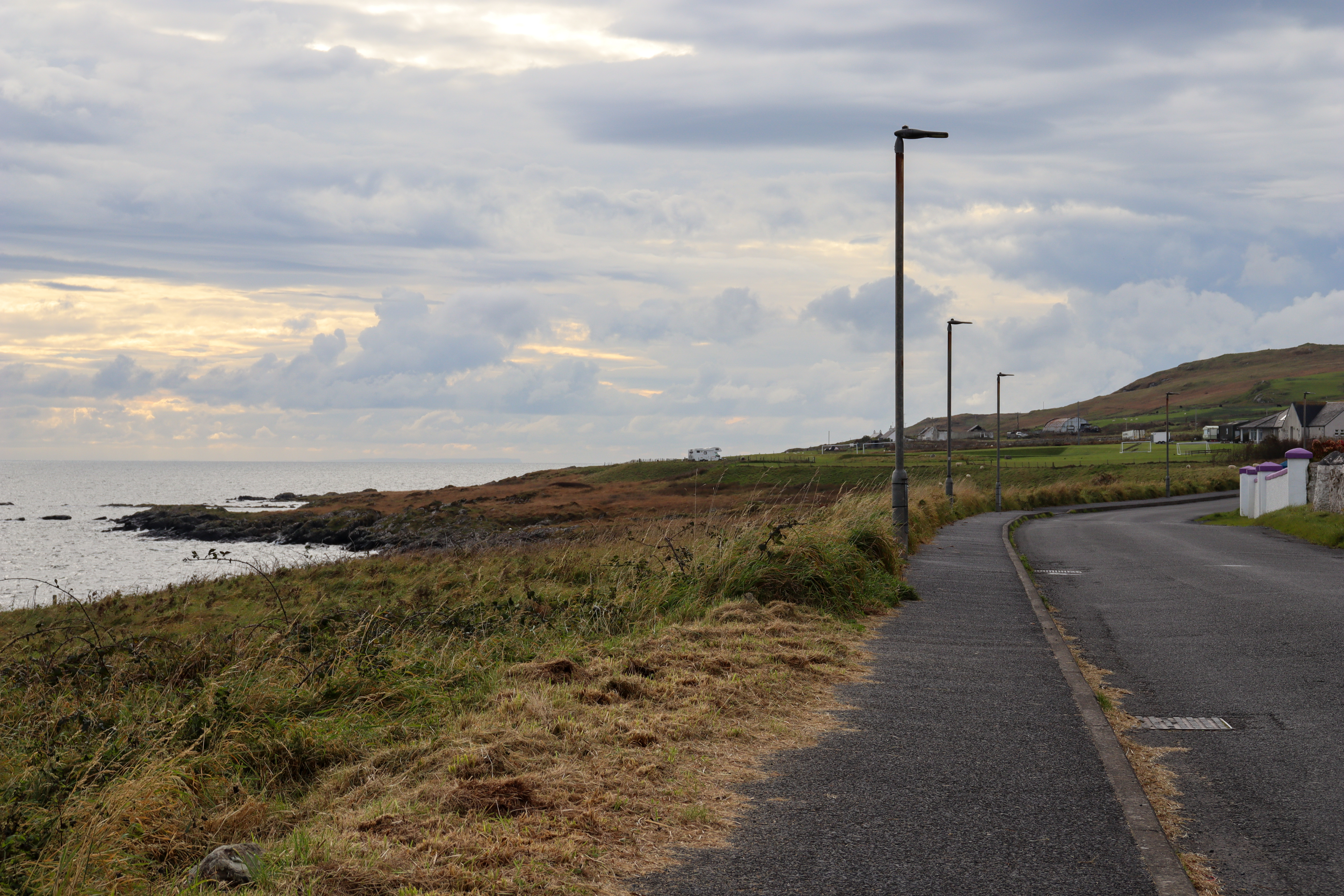 Narrow pavement along one side of a country road. Our motorhome is visible on a hill in the far distance.