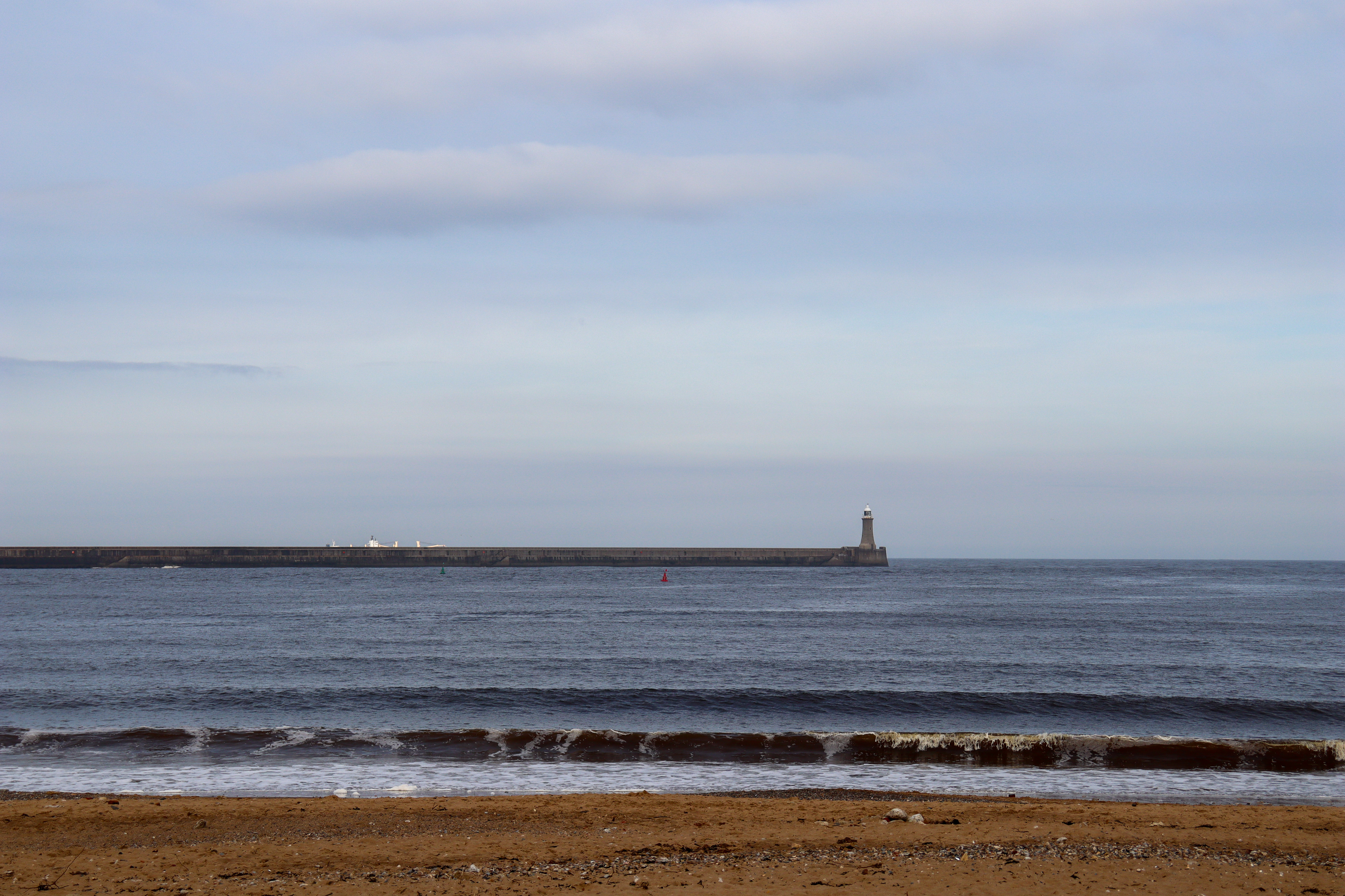 Lighthouse at the end of a pier stretching out to sea. The top of a ferry is just in view behind the pier.